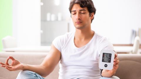 Man macht auf dem Sofa und Yoga und hält ein Blutdruckmessgerät in der Hand