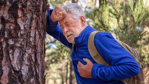 Männlicher, älterer Wanderer mit Herzbeschwerden lehnt sich an Baum 